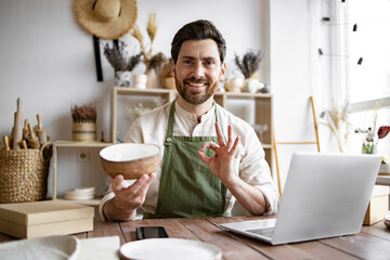 Happy worker in apron holds unique bowl created in personal style against background of shelves...