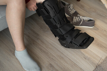 Injured woman with black splint on her leg sits on sofa at home. Ankle brace close-up