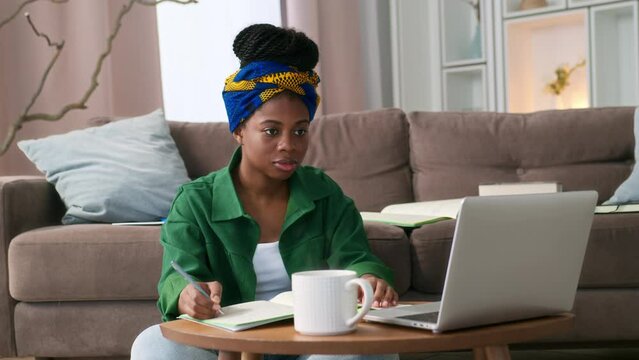 Young black woman with braids sitting on floor in living room at home, working on laptop and notebooks to take notes. African American female student at online course or freelancer working remotely.