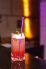 Close-up of a strawberry alcoholic cocktail in a glass with foam decorated with a sprig of rosemary and a straw stands on the bar counter in a beautiful modern bar, copy space. Vertical photo