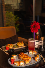 Set of various delicious mouth-watering sushi rolls on the table with a cocktail, soy sauce, chopsticks and a vase with a flower in the restaurant in the evening, a selective focus. Vertical photo