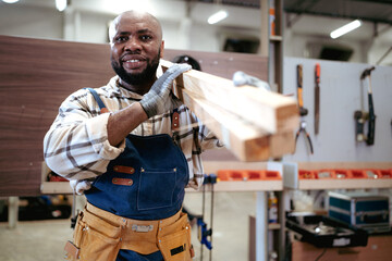 Male carpenter carrying wood planks on his shoulder. Multiracial woodworker wearing safety apron...
