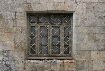 square frame window with a forged metal grill against a wall of beige stone blocks. one window behind  iron bars on a gray marble wall against the sidewalk outside.