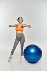 A pregnant woman in activewear stands next to a bright blue exercise ball on a grey background.