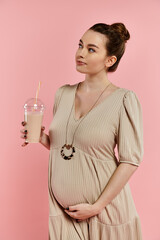 A pregnant woman in a dress joyfully holding milkshake against a pink background.