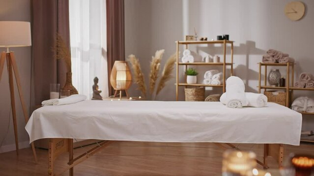 In spa salon with soothing and serene atmosphere. On table laying candles and sounding tranquil music. In treatment room located massage bed with soft linens. Stylish interior concept.