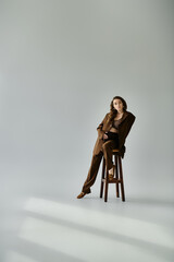 Young pregnant woman in brown suit gracefully sits atop a wooden chair against a grey background.