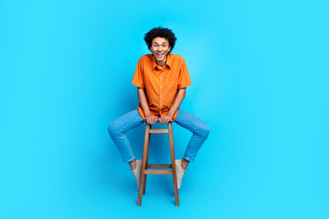 Full length photo of attractive funny guy wear orange shirt sitting chair having fun isolated blue...
