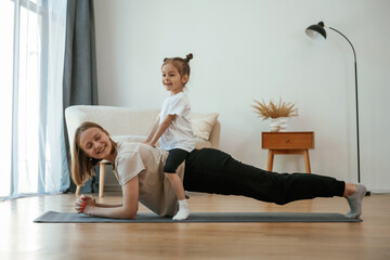 Obraz premium Kid is sitting on a back of woman. Young mother with little girl are doing yoga at home