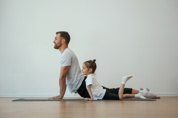 Against white wall, stretching. Father with little daughter are doing yoga at home