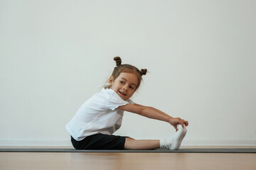 Fototapeta premium Side view, stretching. Cute little girl is sitting on the yoga mat and doing exercises
