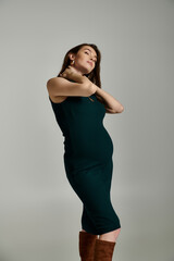 A young pregnant woman elegantly stands in a green dress, exuding grace and charm.