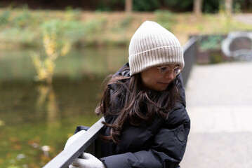 A small beautiful sad girl in glasses and a hat stands near the handrails of a pond in late autumn...