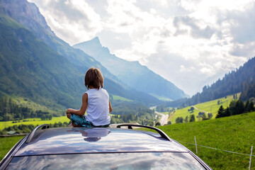 Cute child, boy sitting on a car ceiling, meditating enjoying the quietness of nature, travel on the road to scenic mountains, summertime