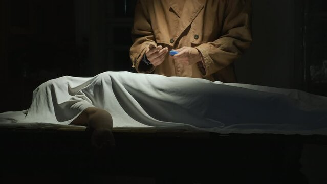 Maniac killer in a cloak stands over a victim covered with a white cloth with a knife in a dark room