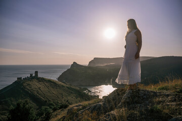 A woman stands on a rocky hill overlooking the ocean. She is wearing a white dress and she is...