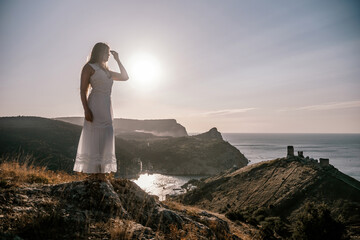 A woman stands on a rocky hill overlooking the ocean. She is wearing a white dress and she is...