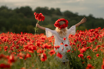 A young girl is standing in a field of red poppies. She is wearing a white dress and a red headband.