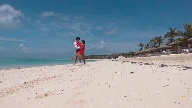 Young couple on ocean shore salsa dancing under shade of passing bird, barefoot girl in red tunic lifting and leaning her knee against her boyfriend thigh, row of white deck chairs under straw awnings