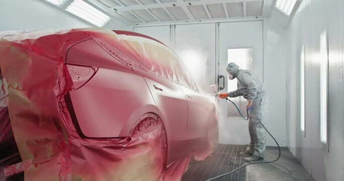 A skilled worker in protective gear spray painting a red car inside a professional automotive paint chamber at a car service station. Worker Spray Painting Red Car in Auto Body Shop