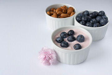 Ramekin with pink yogurt on a white background. Spoon in yogurt. Blueberries with yogurt. Almond nuts. Pink carnation flower. Copy space. Close-up, macro