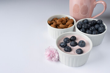 Ramekin with pink yogurt on a white background. Blueberries with yogurt. Almond nuts. Pink carnation flower. Pink cup in the background. Copy space. Close-up, macro