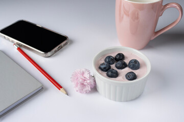 Rose yogurt ramekin is decorated with blueberries. Pink carnation flower. There is a phone, a notepad and a pencil in the background. Pink cup. Close-up. Copy space. Breakfast in the office