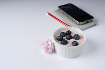 Rose yogurt ramekin is decorated with blueberries. Pink carnation flower. There is a phone, a notepad and a pencil in the background. Close-up. Copy space. Breakfast in the office