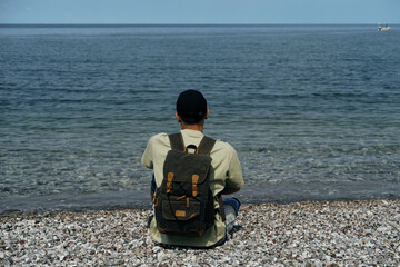 Travel, people and holiday concept. A young man with a retro backpack sits on the pebbly shore of...