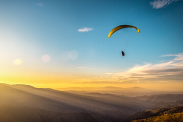 Paraglider silhouette flying over misty mountain valley in beautiful warm sunset colors