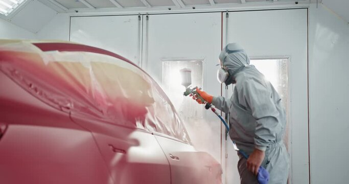 Professional car painter wearing protective gear, applying red paint to a car in an auto service chamber. Concept of automotive paint services and car body repair. Car manufacturing factory