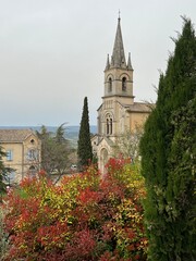 Naklejka premium A church steeple juts into the sky in a small village in Provence, France on a cloudy day with colourful bushes and trees in the foreground