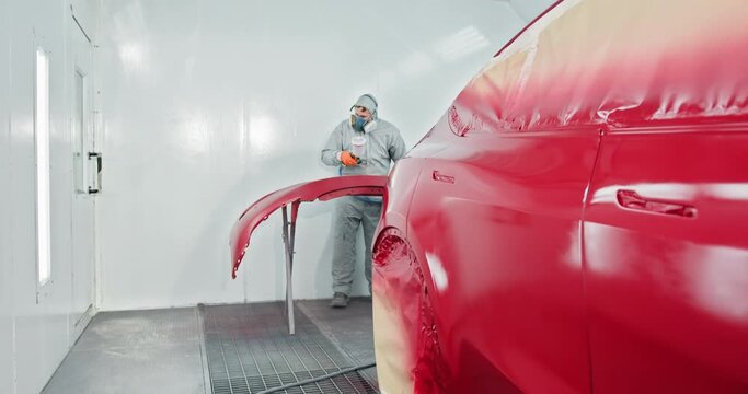 A car painter wearing protective gear is applying red paint to an automobile bumper in a service chamber. Automotive paint services and bodywork repair in progress. Car manufacturing factory
