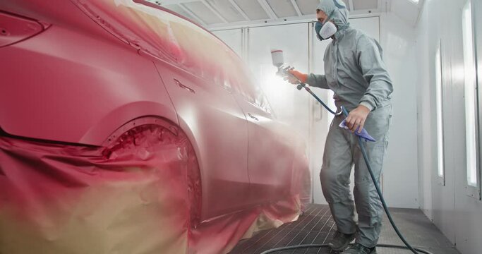 A car painter in a protective suit and mask is spray painting a car in a specialized booth in an auto body shop. Auto Painter Applying Paint in Spray Booth. Concept of automotive paint services