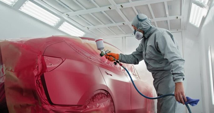 A car painter in a protective suit and mask is spray painting a car in a specialized booth in an auto body shop. Auto Painter Applying Paint in Spray Booth. Concept of automotive and car body repair