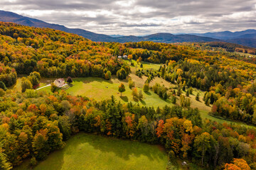 Autumn forest adorns isolated farmhouse under clouds, mountain backdrop. Dron view. Vermont, USA.