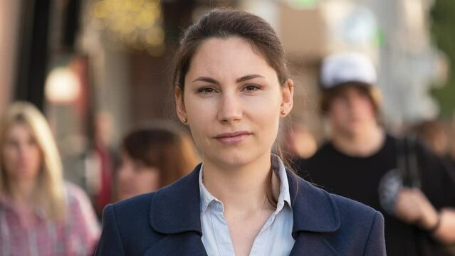 A woman in a formal jacket and shirt stands in the middle of the street and looks at the camera. A crowd of people walks around her. Daily bustle concept