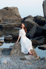 Serene woman in white dress sitting on rocky ocean shore with arms outstretched in peaceful...