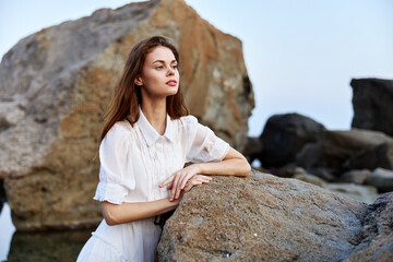 Confident woman sitting on rocky ocean shore with hands on hips in white shirt on a sunny day