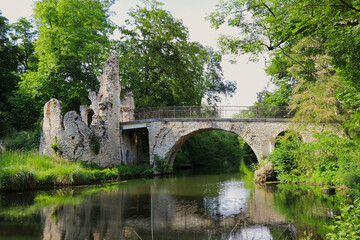 Fototapeta premium an ancient stone bridge and ruins over a tranquil river, surrounded by lush green trees. The scene combines historical charm with natural beauty, evoking a peaceful ambiance.