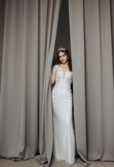Fototapeta premium A woman in a white dress stands behind a curtain, looking out into the room. The curtain is drawn, creating a sense of mystery and intrigue. The woman's elegant attire