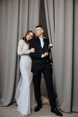 A bride and groom are posing for a picture in front of a curtain. The bride is wearing a tiara and...