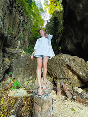 Naklejka premium A woman in an oversized white t-shirt and shorts stands on a tree stump in a lush forest, surrounded by tall cliffs and greenery, looking serene and contemplative.