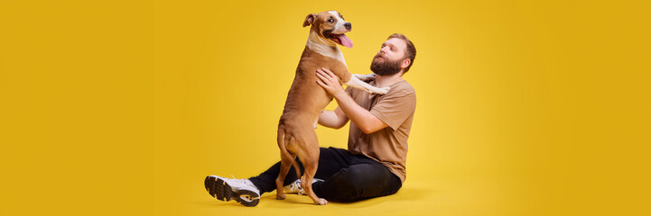 Banner. Man in casual attire sits on floor happily hugging his dog, who stands on its hind leg...