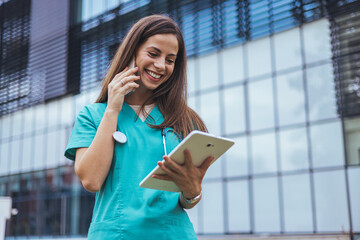 A cheerful Caucasian female nurse in teal scrubs actively engages with a tablet outside a medical...