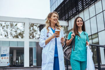 Two cheerful Caucasian women in healthcare attire, a doctor and nurse, walk and converse outside a...