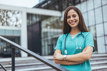 A smiling Caucasian healthcare worker stands confidently outdoors with arms crossed, wearing...