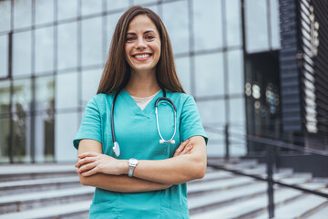 A smiling Caucasian woman in teal scrubs stands confidently with arms crossed outside a medical...