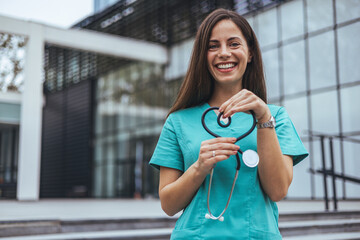 A joyful Caucasian female nurse forms a heart with her stethoscope, displaying care outside a...