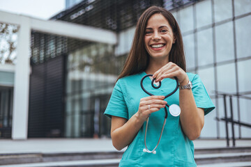 A joyful Caucasian female nurse forms a heart with her stethoscope, displaying care outside a...